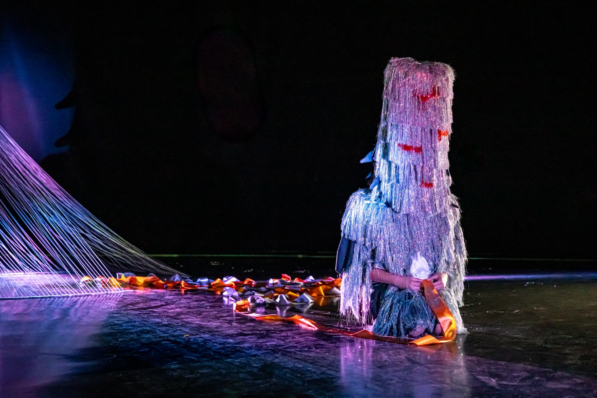 Une personne assise sur scène, vêtue d’un costume texturé et scintillant, portant un masque couvrant le visage, entourée de rubans colorés et d’un faisceau lumineux violet en arrière-plan.